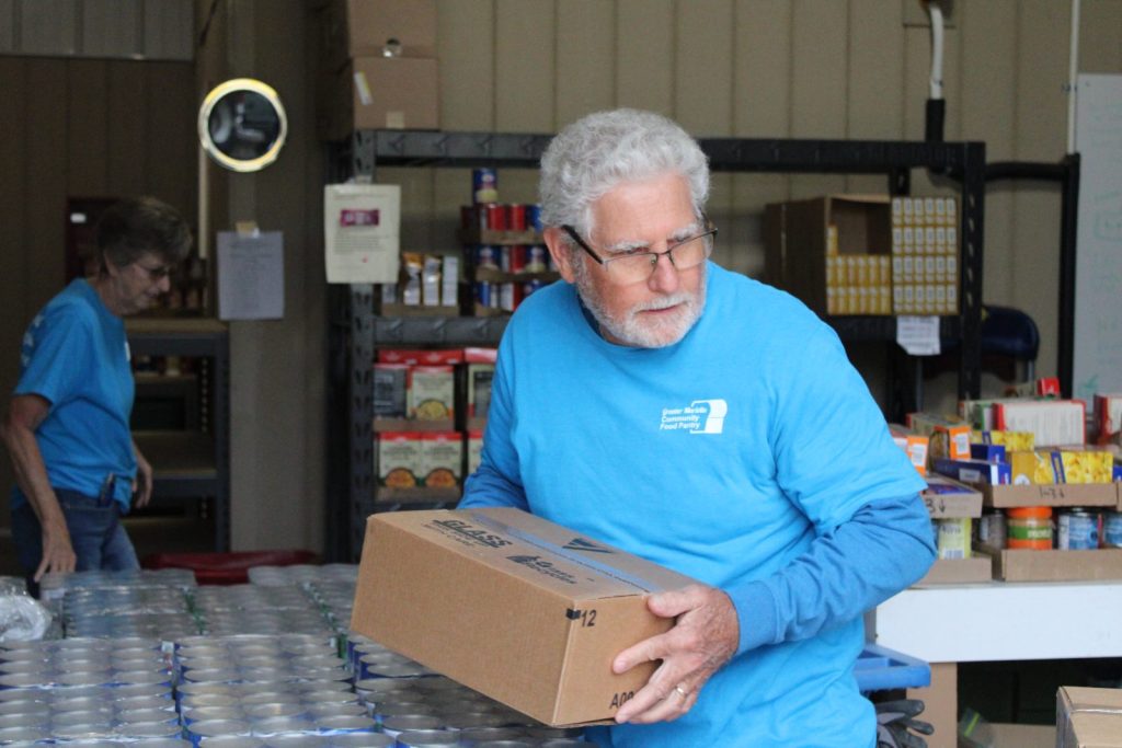 Photo of an older man volunteer holding a box of shelf stable food inside the GMFP.