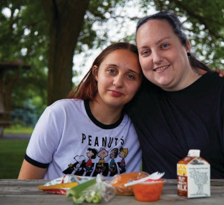 photo of a mom and her teen daughter with their arms around each other. They are sitting outside with a summer meals lunch in front of them.