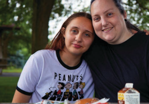 photo of a mom and her teen daughter with their arms around each other. They are sitting outside with a summer meals lunch in front of them.